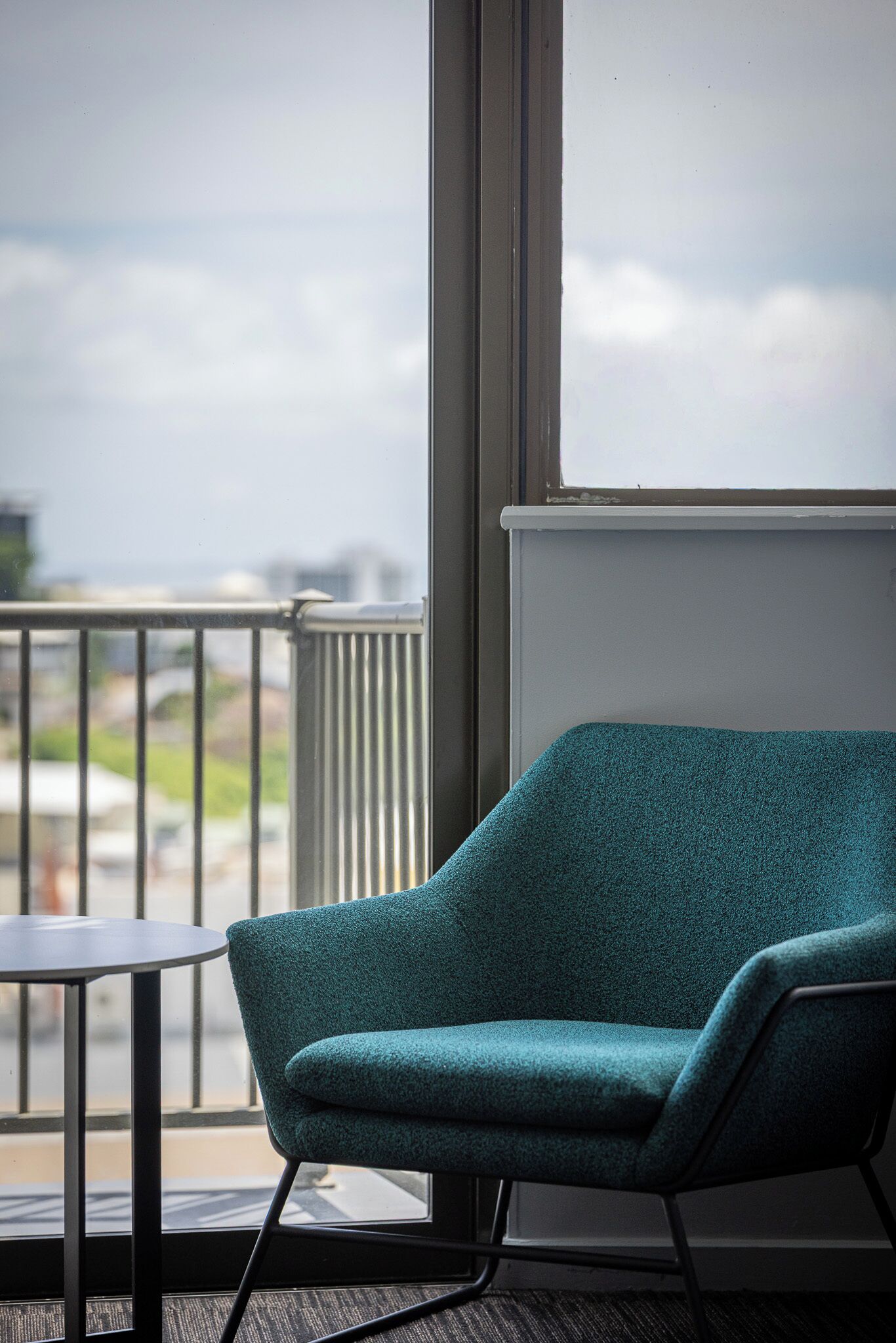 A teal armchair and a small table on a balcony with a view of buildings and greenery.