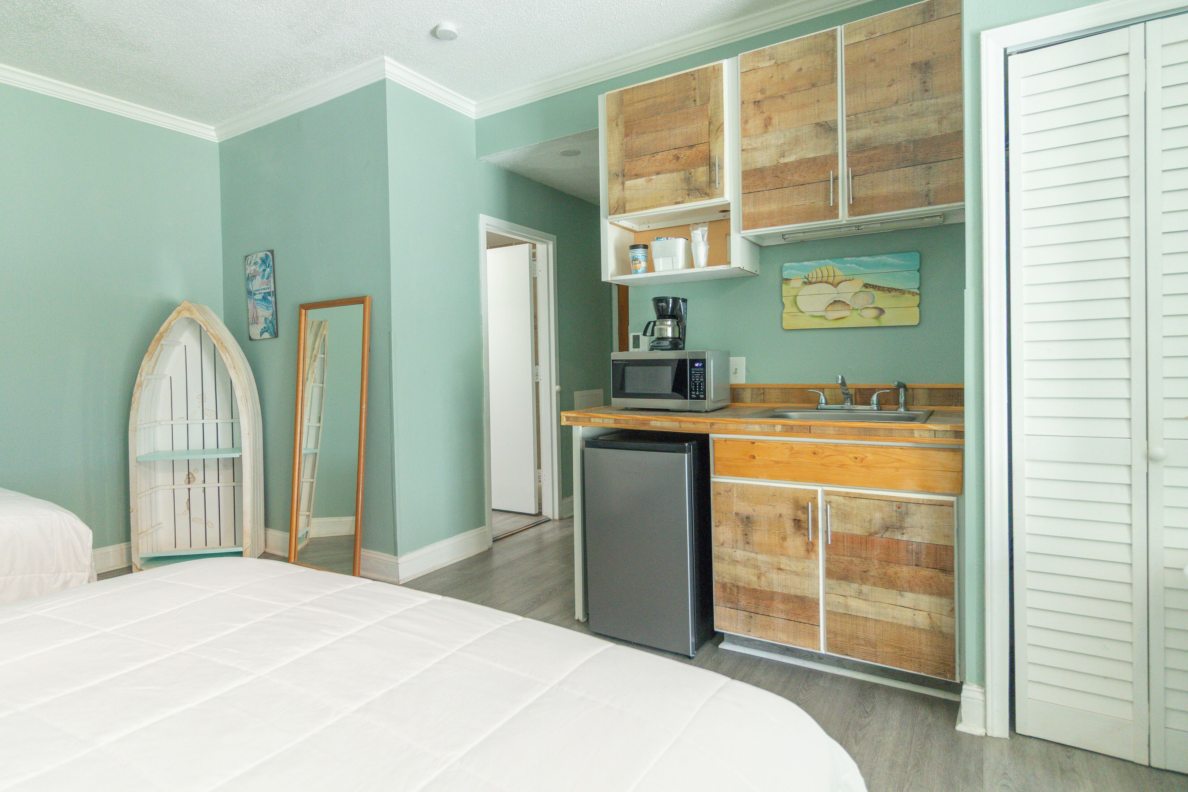 A bedroom with a bed, a small kitchenette with a microwave, and a refrigerator. There is a wooden cabinet above the sink and a door with a mirror.