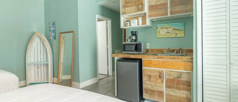 Un dormitorio con cama, una kitchenette pequeña con microondas y un refrigerador. Hay un mueble de madera sobre el lavamanos y una puerta con espejo.
