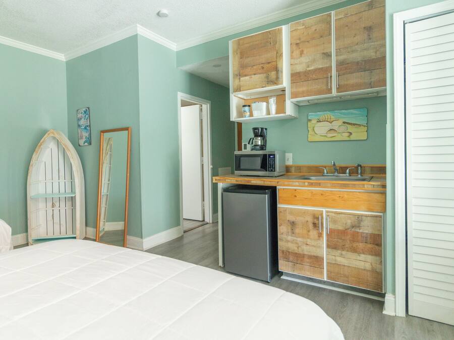 A bedroom with a bed, a small kitchenette with a microwave, and a refrigerator. There is a wooden cabinet above the sink and a door with a mirror.