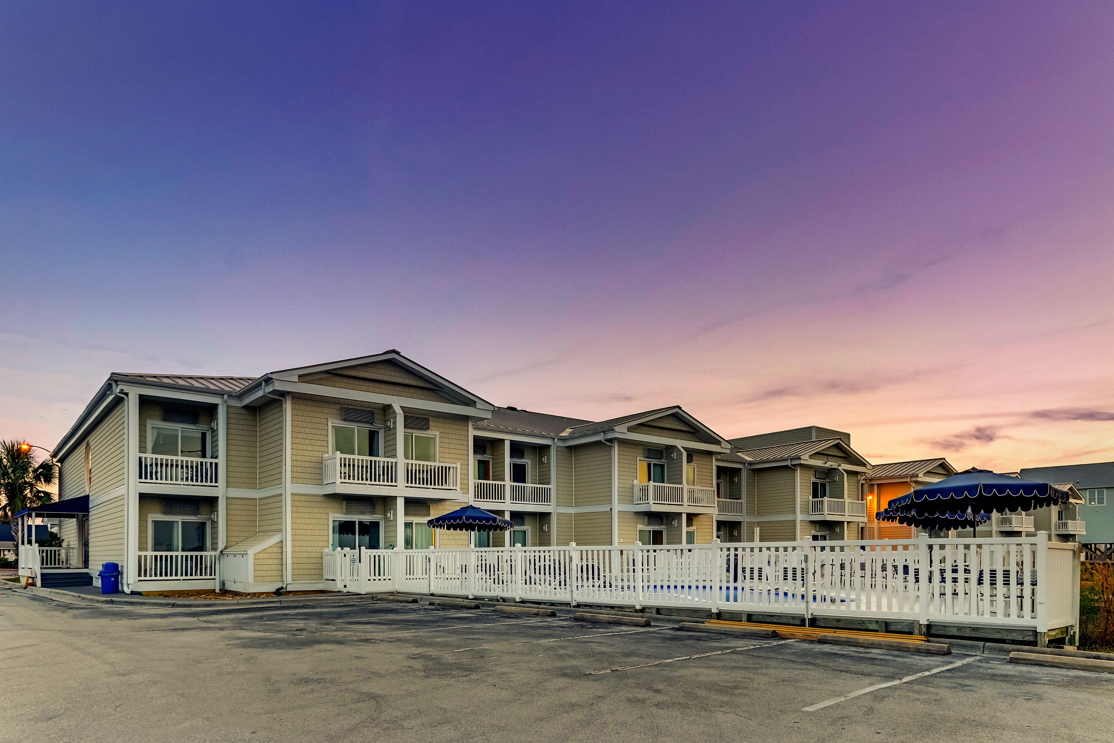A multi-story motel with balconies, a fenced parking area, and a sunset sky.