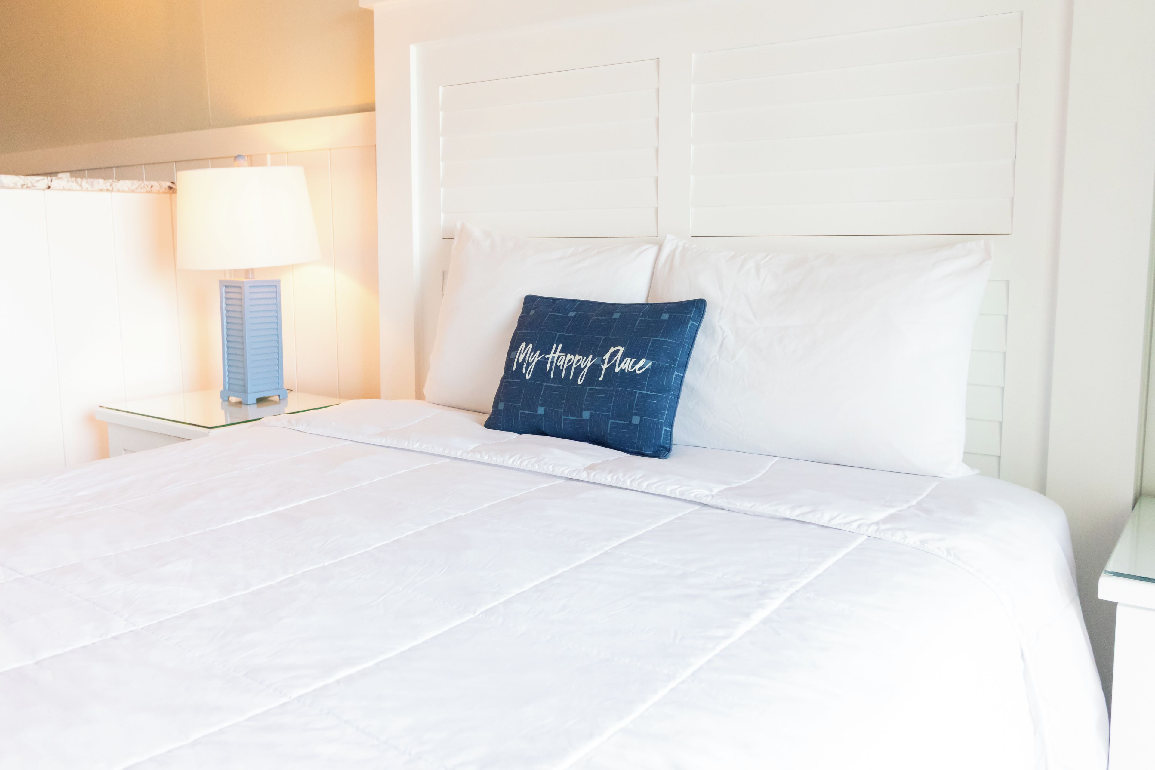 A neatly made bed with white bedding and a single blue pillow with text, a bedside lamp, and a white headboard.