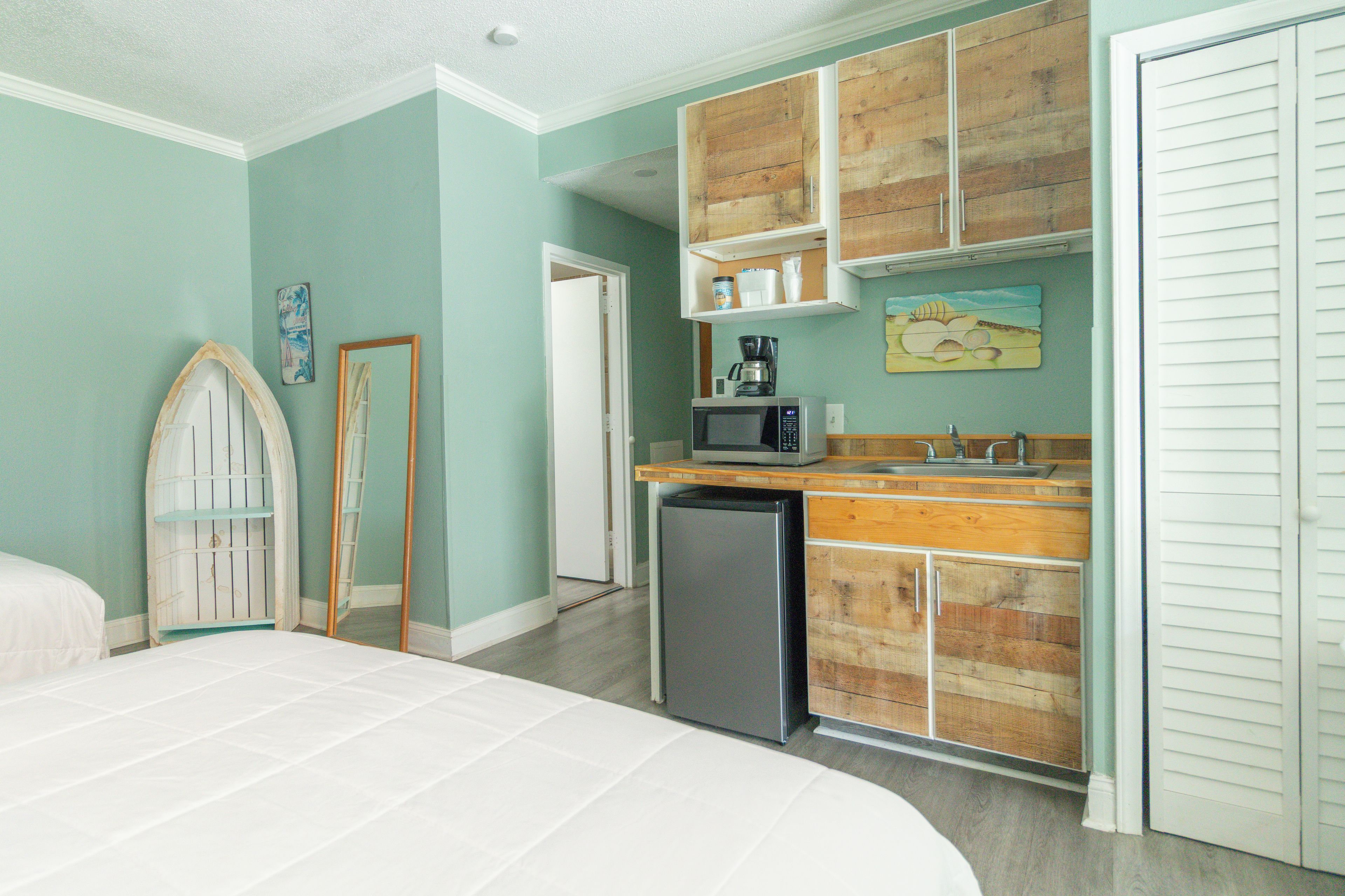 Un dormitorio con cama, una kitchenette pequeña con microondas y un refrigerador. Hay un mueble de madera sobre el lavamanos y una puerta con espejo.