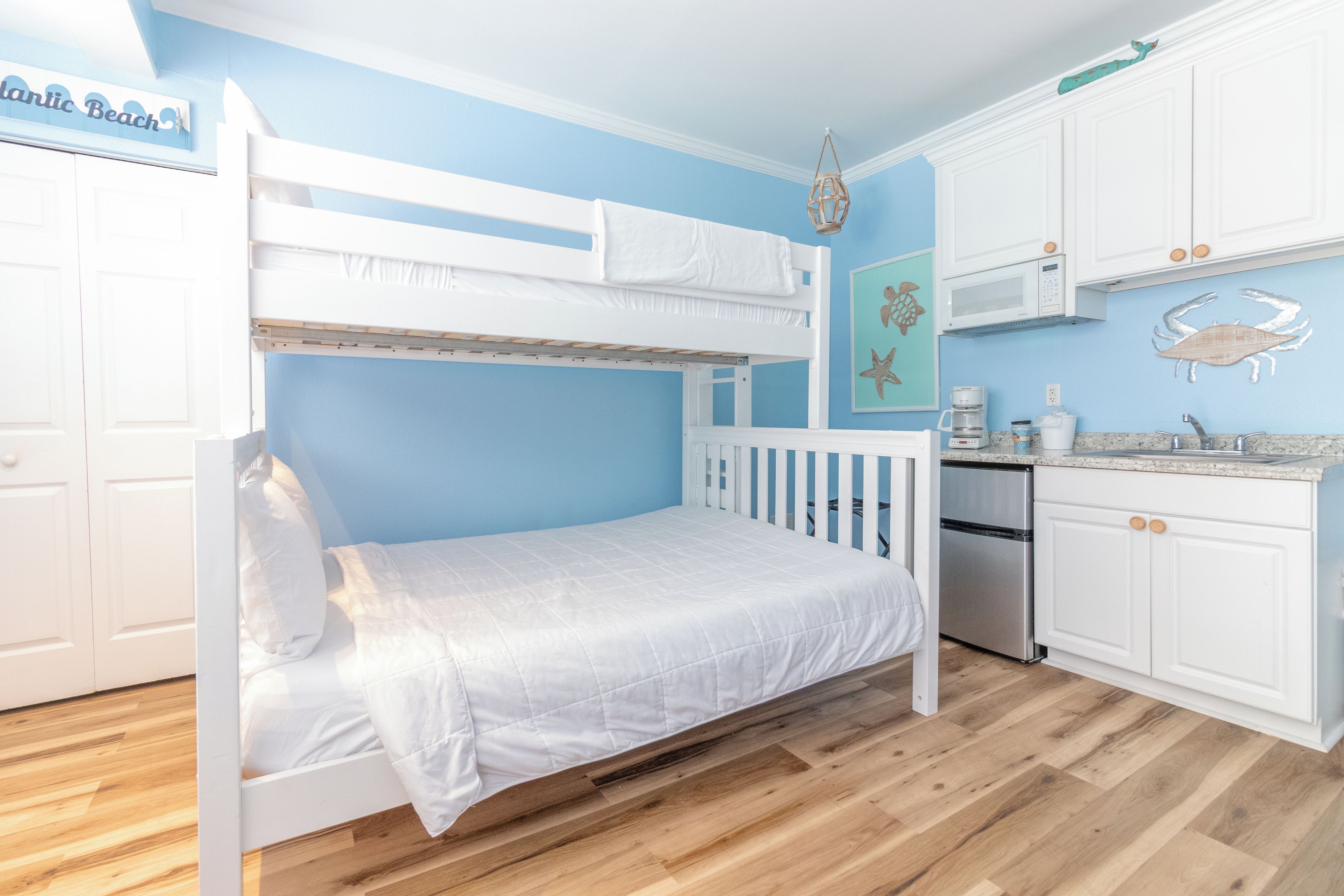 A bunk bed with a white mattress and bedding, a kitchen area with white cabinets and a stainless steel refrigerator, and a blue wall with a decorative fish mural.