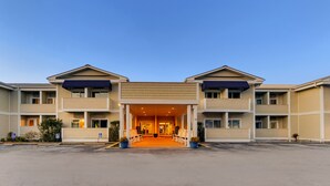 A multi-story hotel with a central entrance, outdoor seating, and a parking lot. - Palm Suites Trademark Collection by Wyndham (Atlantic Beach)