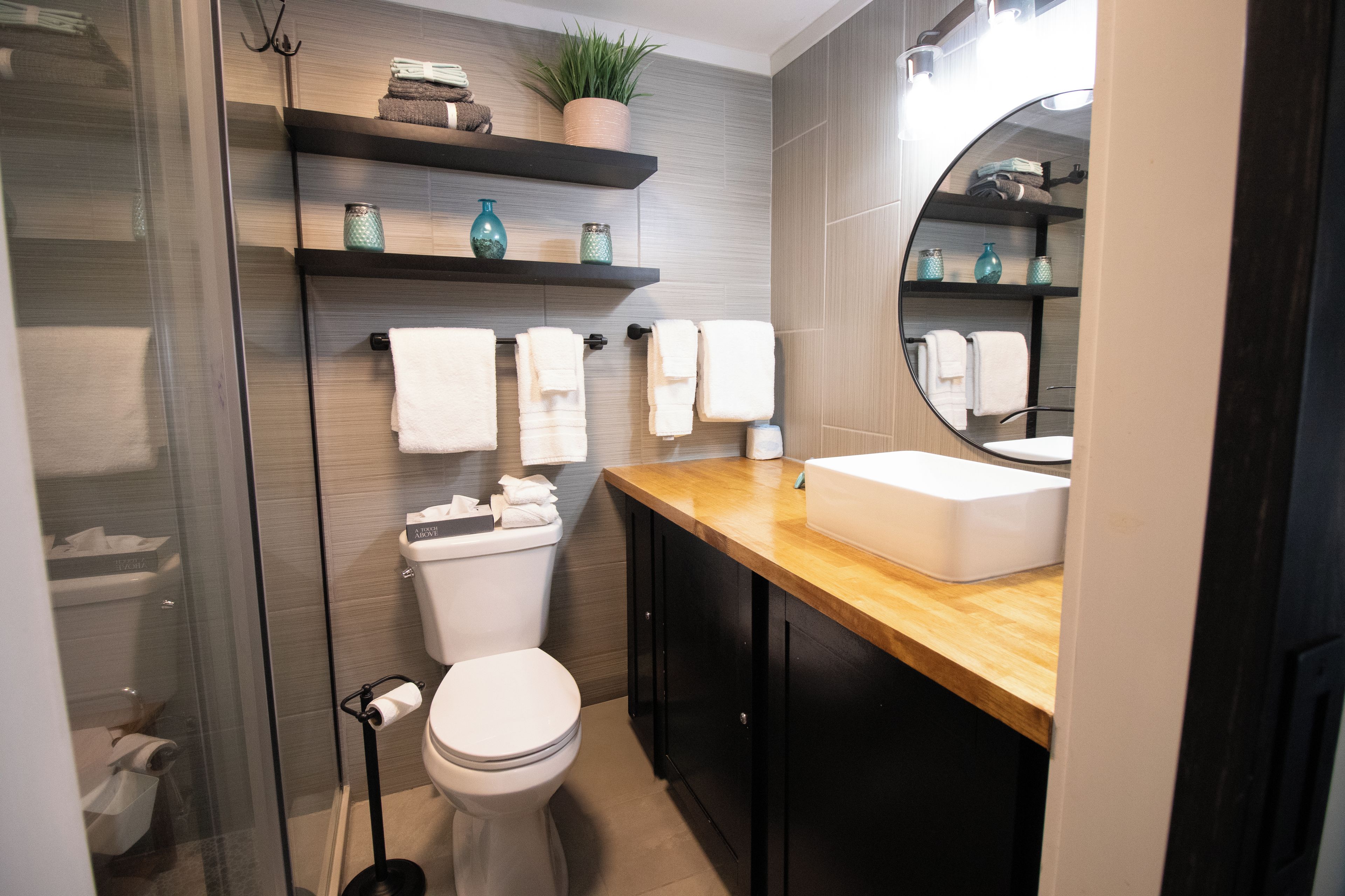 A modern bathroom with a toilet, sink, and mirror. There are shelves with towels and decorative items.
