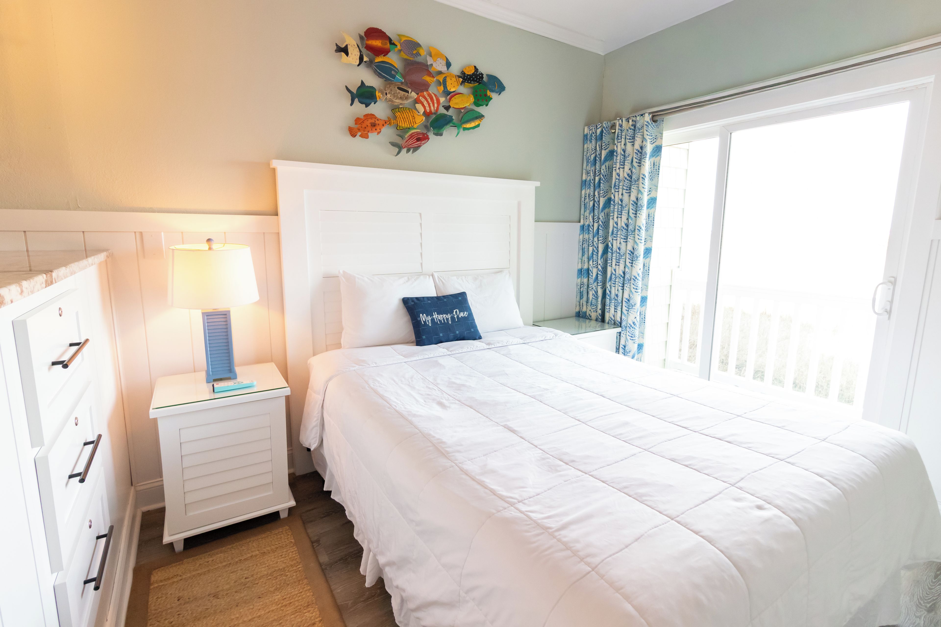 A bedroom with a bed, a nightstand with a lamp, a dresser, and a wall decoration.