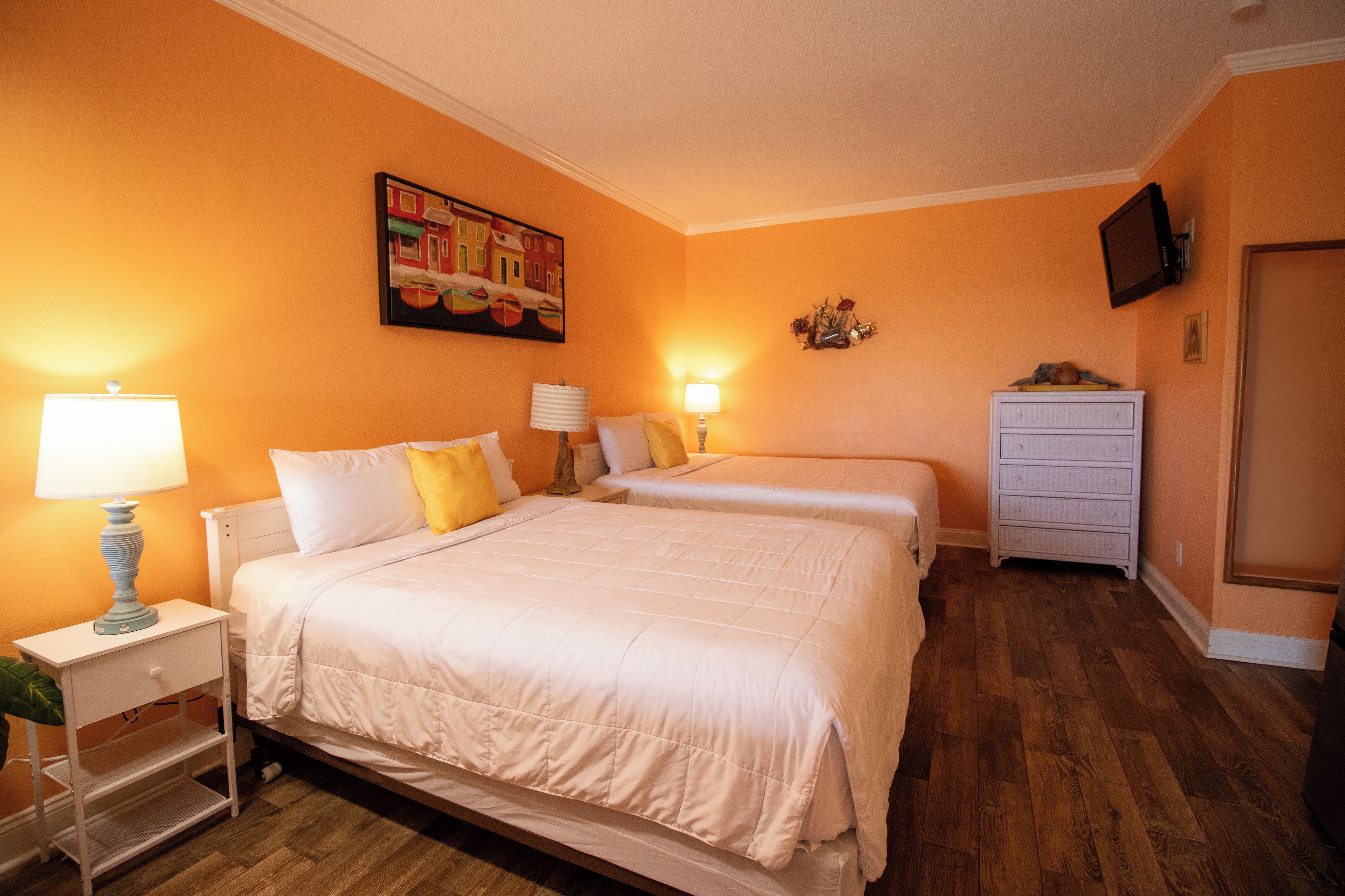 A hotel room with two beds, a dresser, a painting on the wall, and a television mounted on the wall.