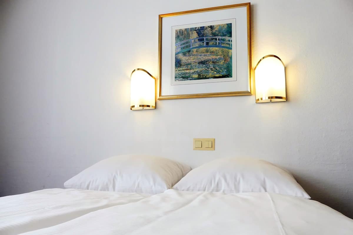 Une chambre d’hôtel avec un lit, deux lampes fixées au mur et un tableau encadré au-dessus du lit.
