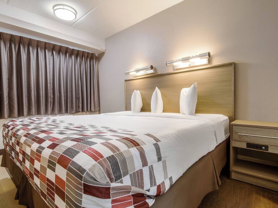 A hotel room with a large bed, a wooden headboard, a nightstand with a drawer, and a window with curtains.