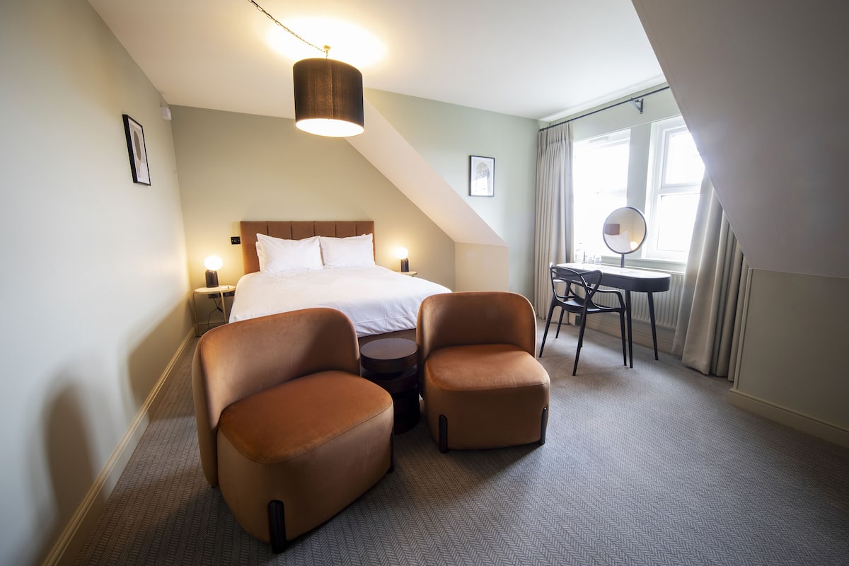 Une chambre d’hôtel comprenant un lit, deux fauteuils, une petite table, un bureau avec une chaise, une lampe et une fenêtre avec des rideaux.