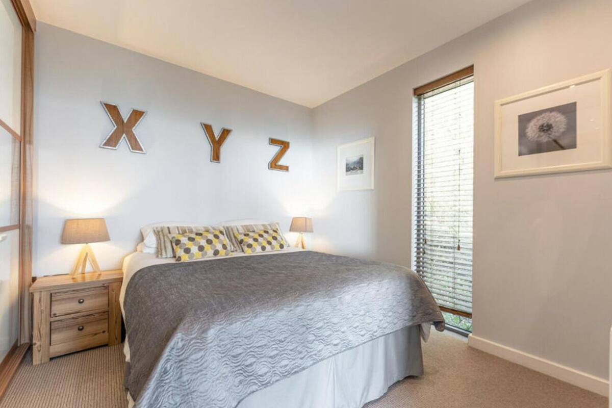 Une chambre à coucher avec un lit, deux tables de chevet, une fenêtre avec des stores et des lettres décoratives fixées au mur.