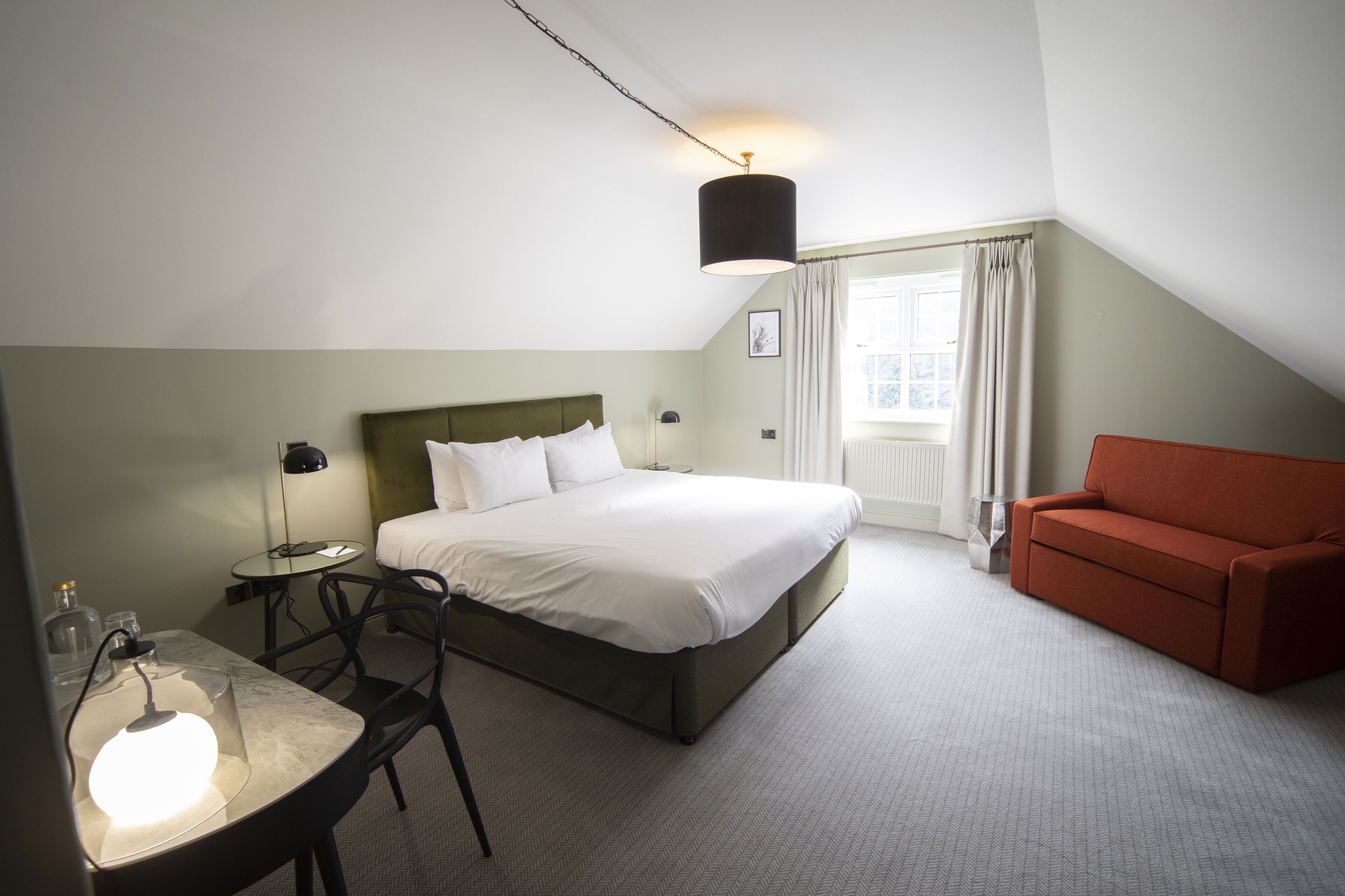A hotel room with a bed, a desk with a lamp, a red sofa, and a window with curtains.