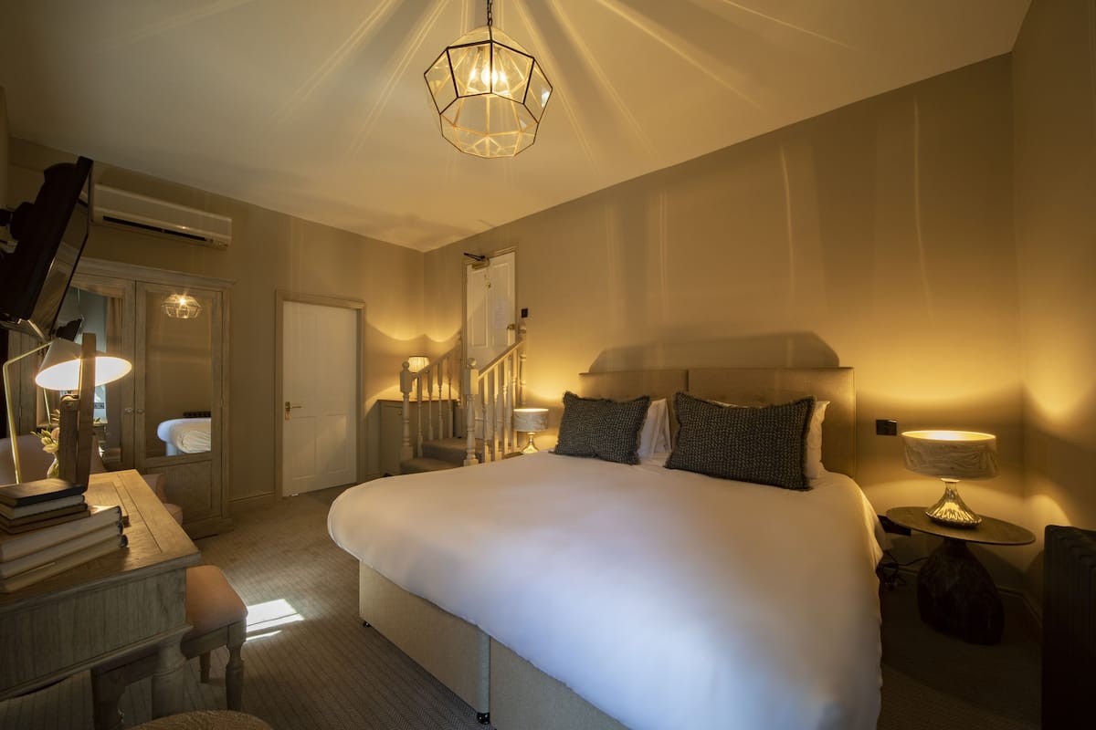 Une chambre d’hôtel avec un grand lit, un bureau avec une lampe, une chaise, une table de chevet avec une lampe, un miroir et un escalier menant à une autre pièce.