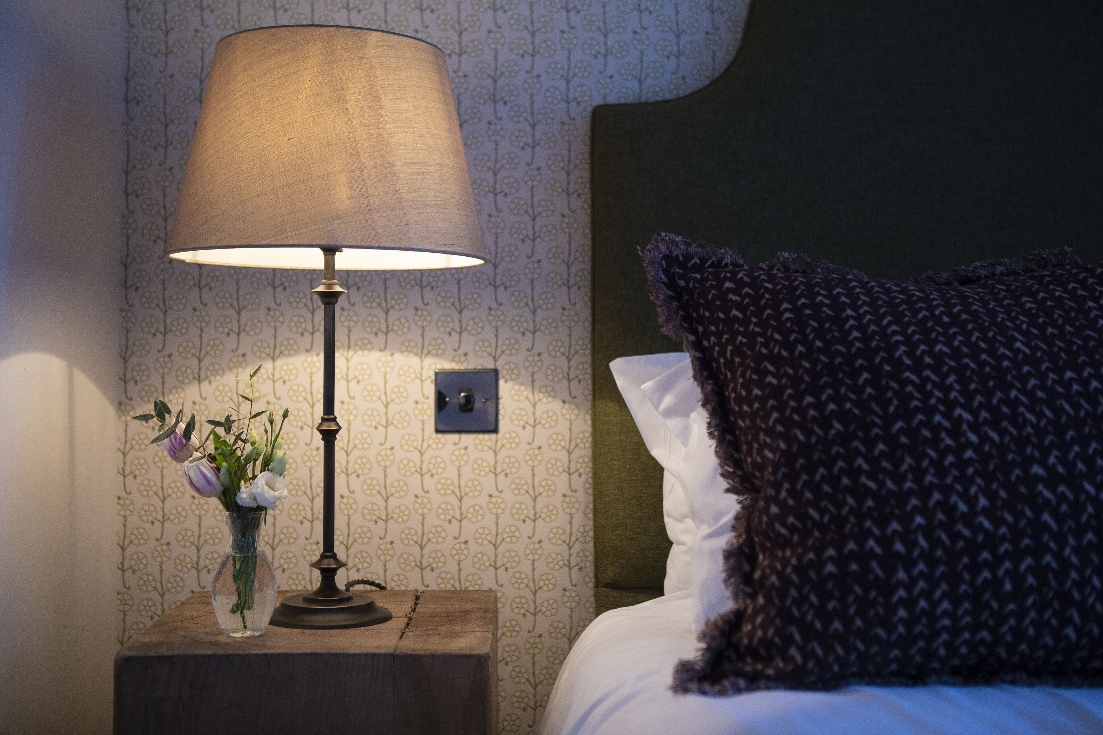 Una cama bien hecha con un cojín oscuro y una lámpara de noche sobre una mesita de madera.