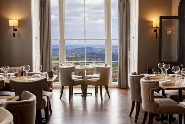 A dining area with a table set for six, large windows with curtains, and a view of the countryside. - The Cottage in the Wood (Malvern)