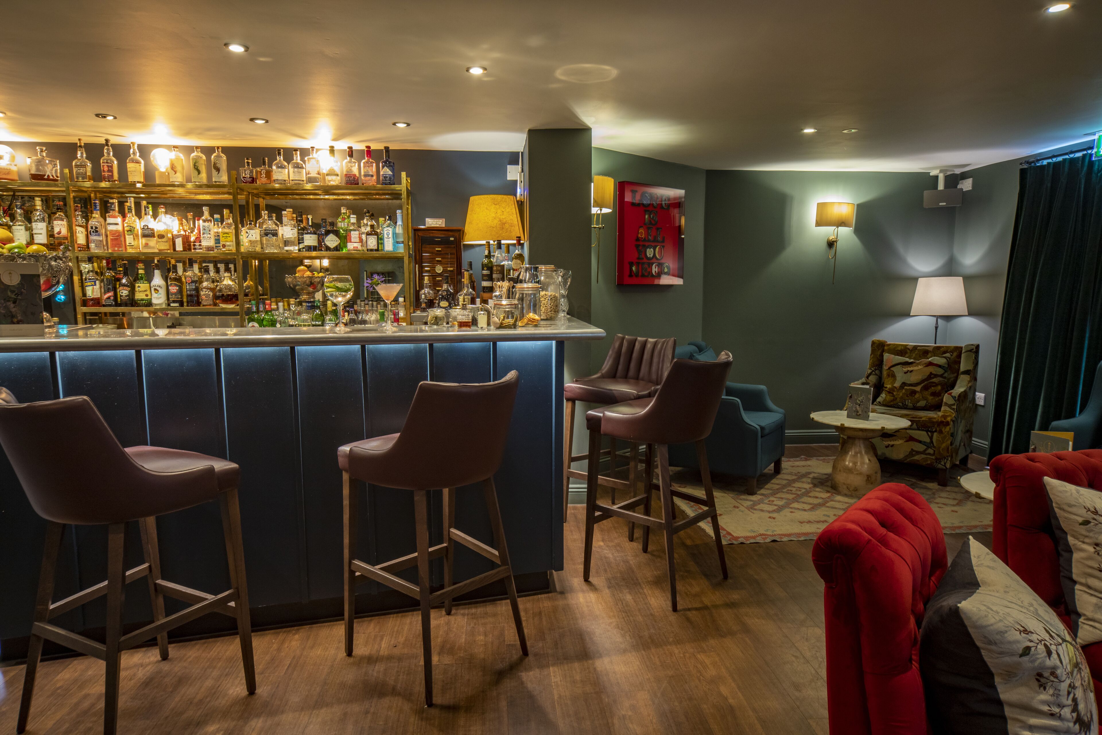 A bar area with stools, a bar counter, and a variety of bottles. There are two armchairs and a red sofa in the seating area.