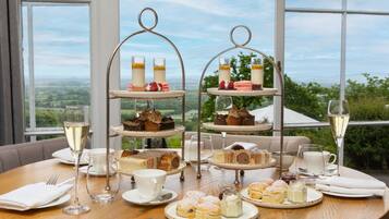 Une table dressée avec une variété de pâtisseries, de gâteaux et de boissons, offrant une vue panoramique à travers de grandes fenêtres.