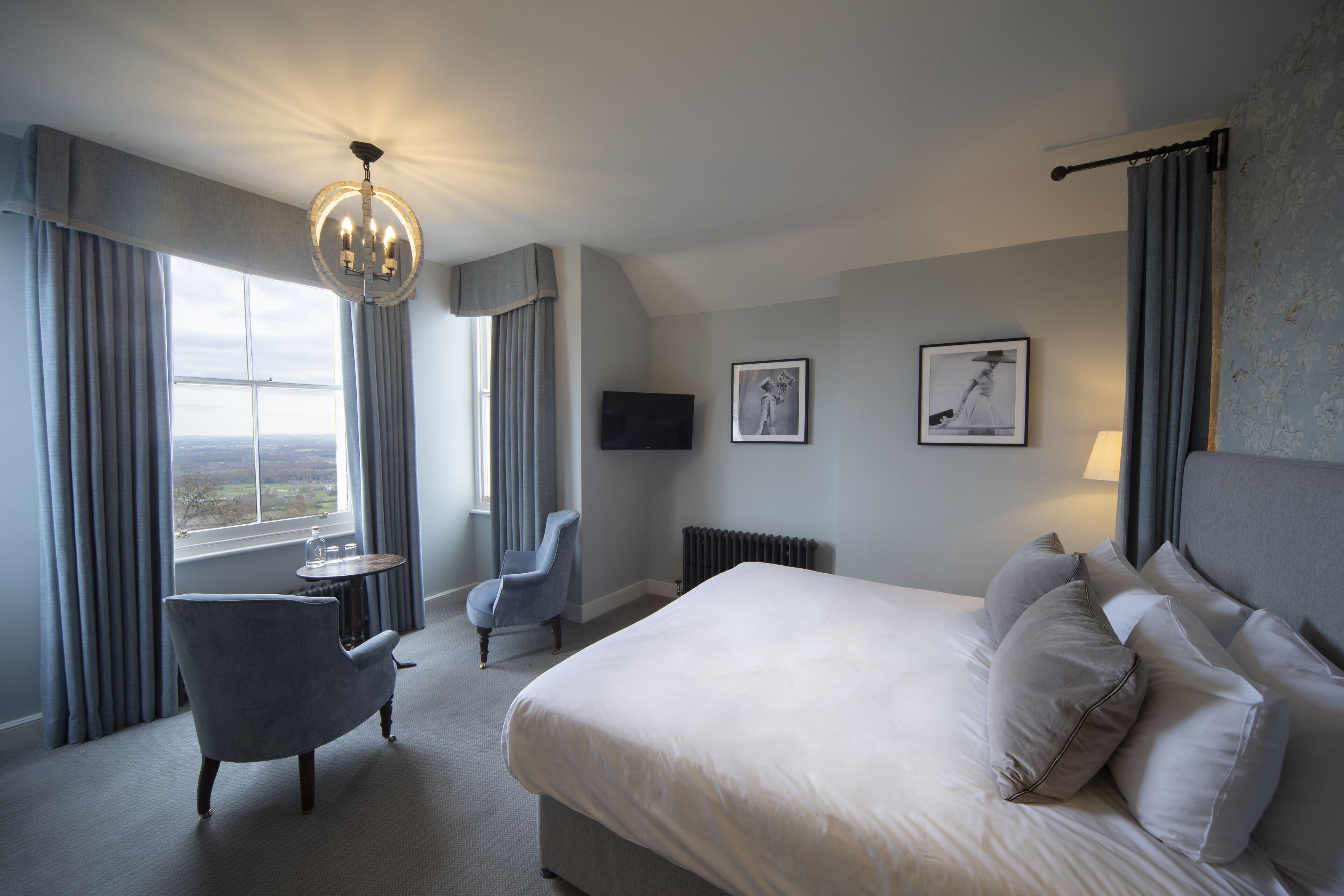 A hotel room with a large bed, a chair, a small table, a chandelier, and a window with curtains.