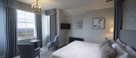 A hotel room with a large bed, a chair, a small table, a chandelier, and a window with curtains.