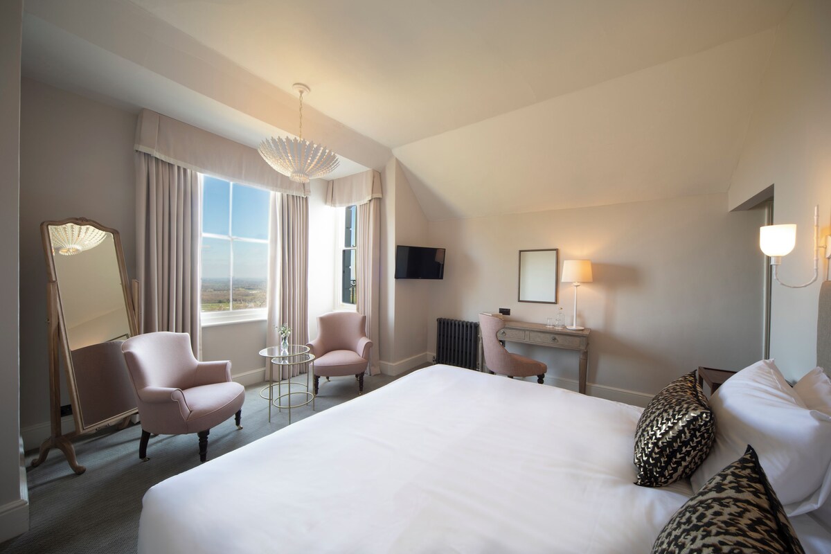 Ett hotellrum med en stor säng, två fåtöljer, ett litet bord, ett skrivbord, en tv och ett fönster med gardiner.