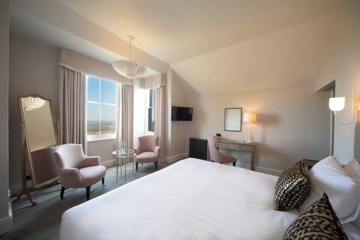 Une chambre d’hôtel avec un grand lit, deux fauteuils, une petite table, un bureau, une télévision et une fenêtre avec des rideaux.