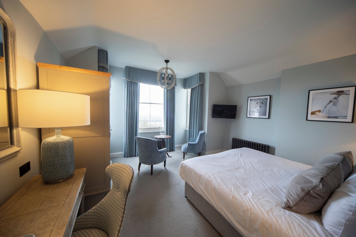 A hotel room with a bed, a desk with a lamp, a chair, a television, and a window with curtains.