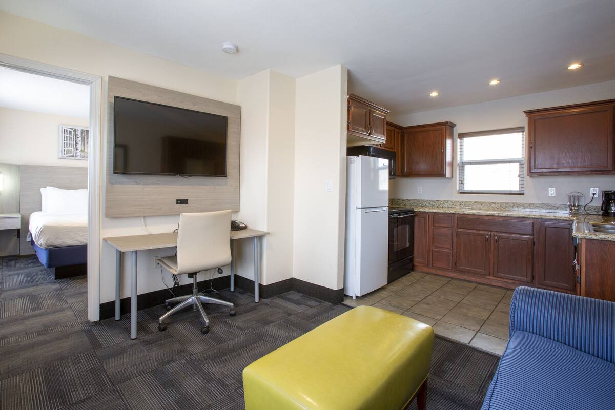Habitación de hotel compacta con kitchenette, cama, escritorio con silla y pequeño rincón de estar con sofá y reposapiés.