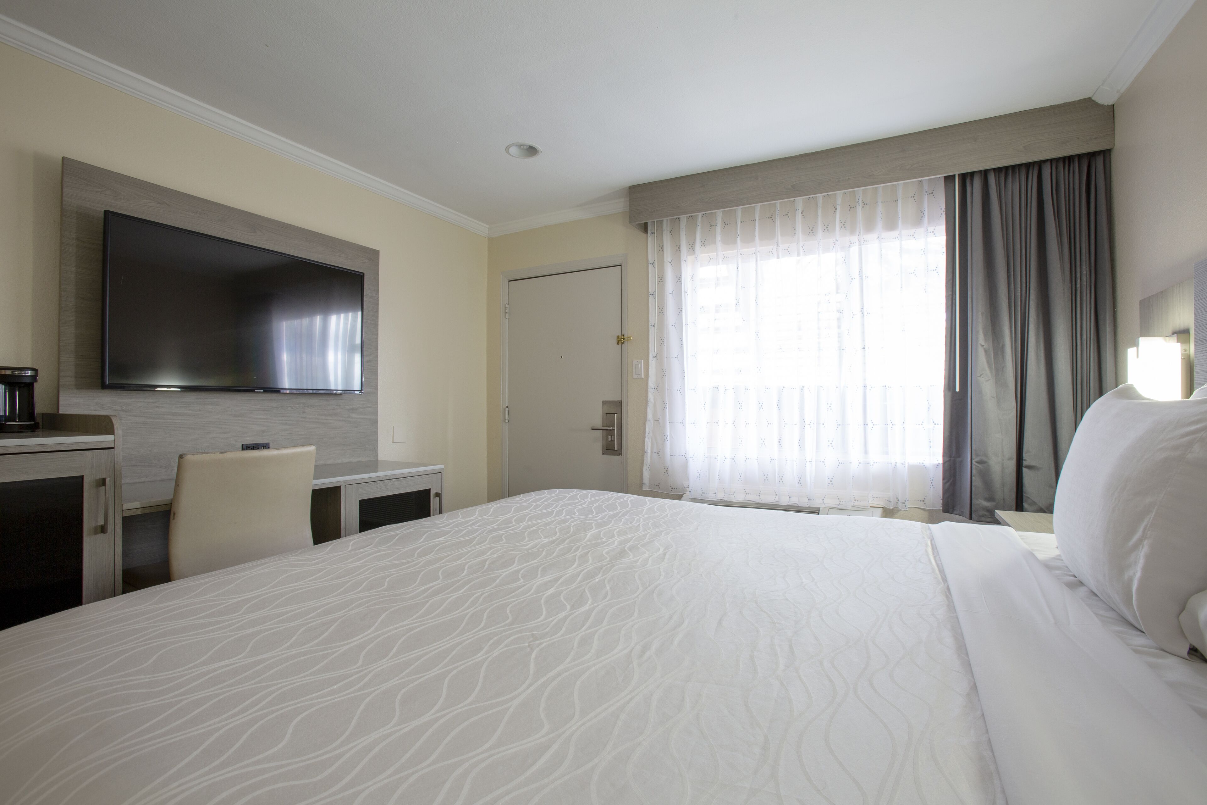 Ein Hotelzimmer mit einem großen Bett, einem flachbildfernseher an der Wand, einem Schreibtisch mit Stuhl und einem Fenster mit Vorhängen.