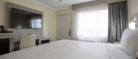 Ein Hotelzimmer mit einem großen Bett, einem flachbildfernseher an der Wand, einem Schreibtisch mit Stuhl und einem Fenster mit Vorhängen.