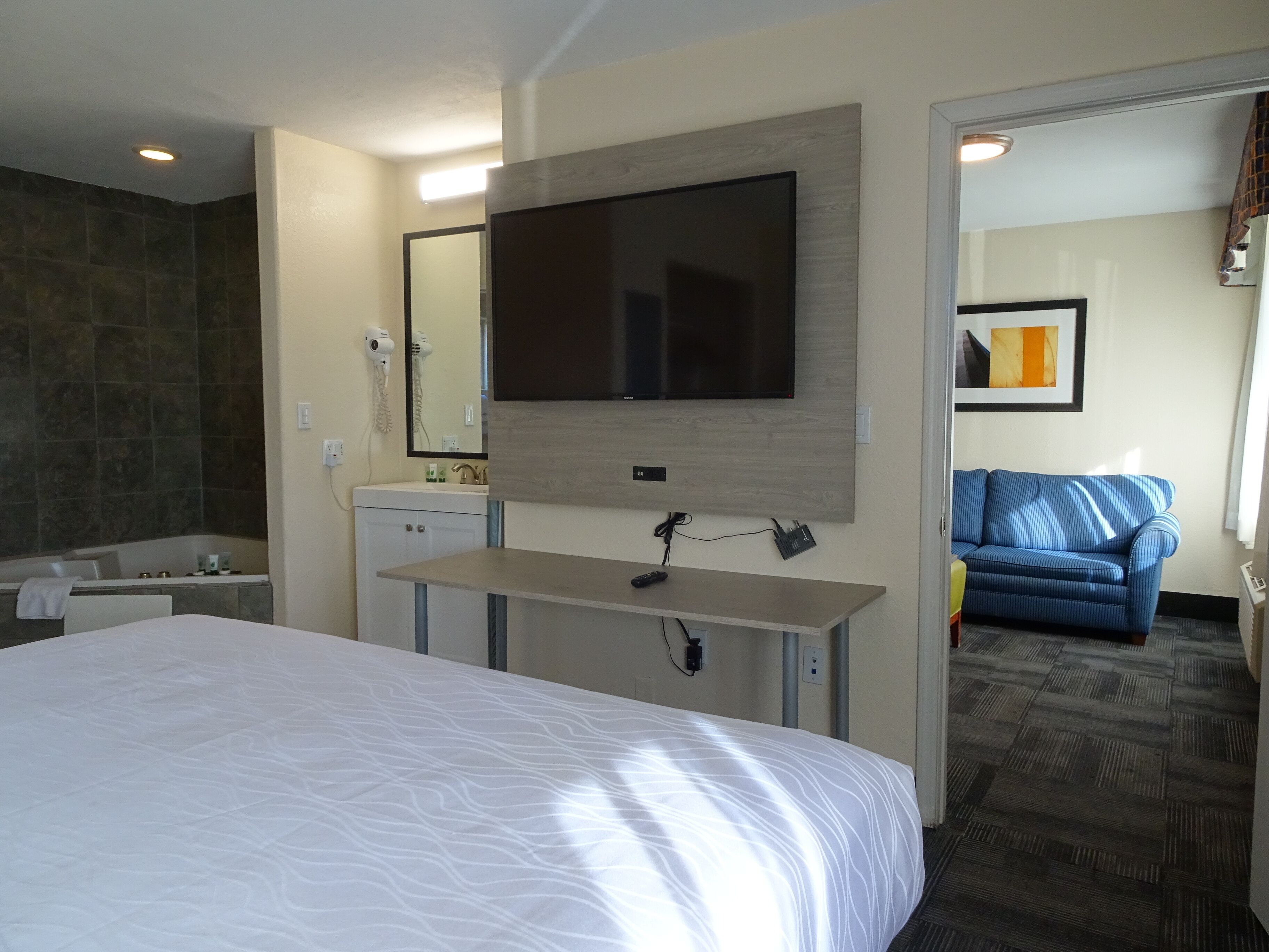 A hotel room with a large bed, a flat-screen TV mounted on the wall, a small desk with a phone, a bathroom with a bathtub, and a blue sofa in the adjacent living area.