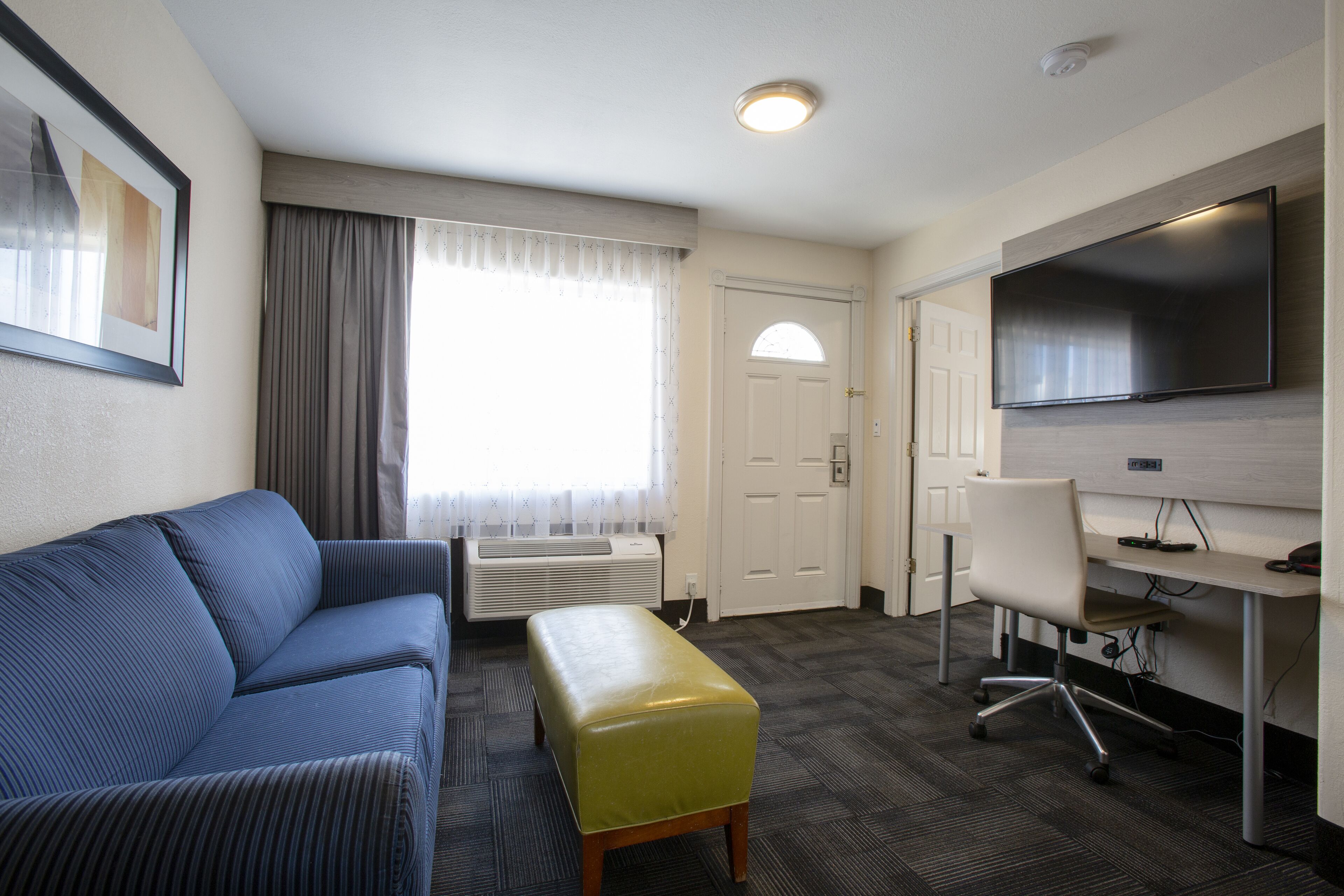 Hotelkamer met een blauwe bank, een geel poefje, een bureau met stoel, een flatscreen televisie en een groot raam met gordijnen.