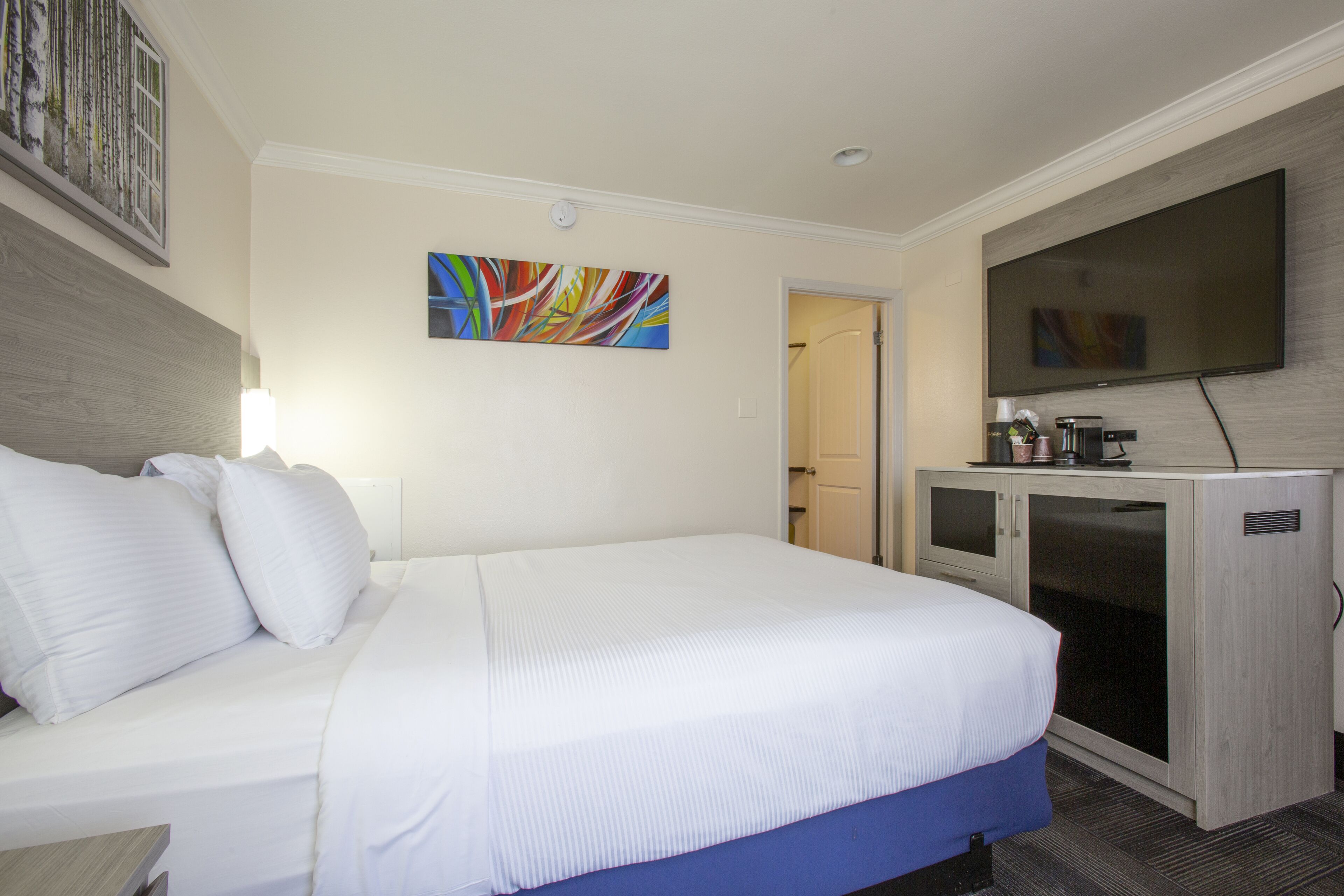 A hotel room with a large bed, a flat-screen TV mounted on the wall, a desk with a lamp, and a coffee machine.