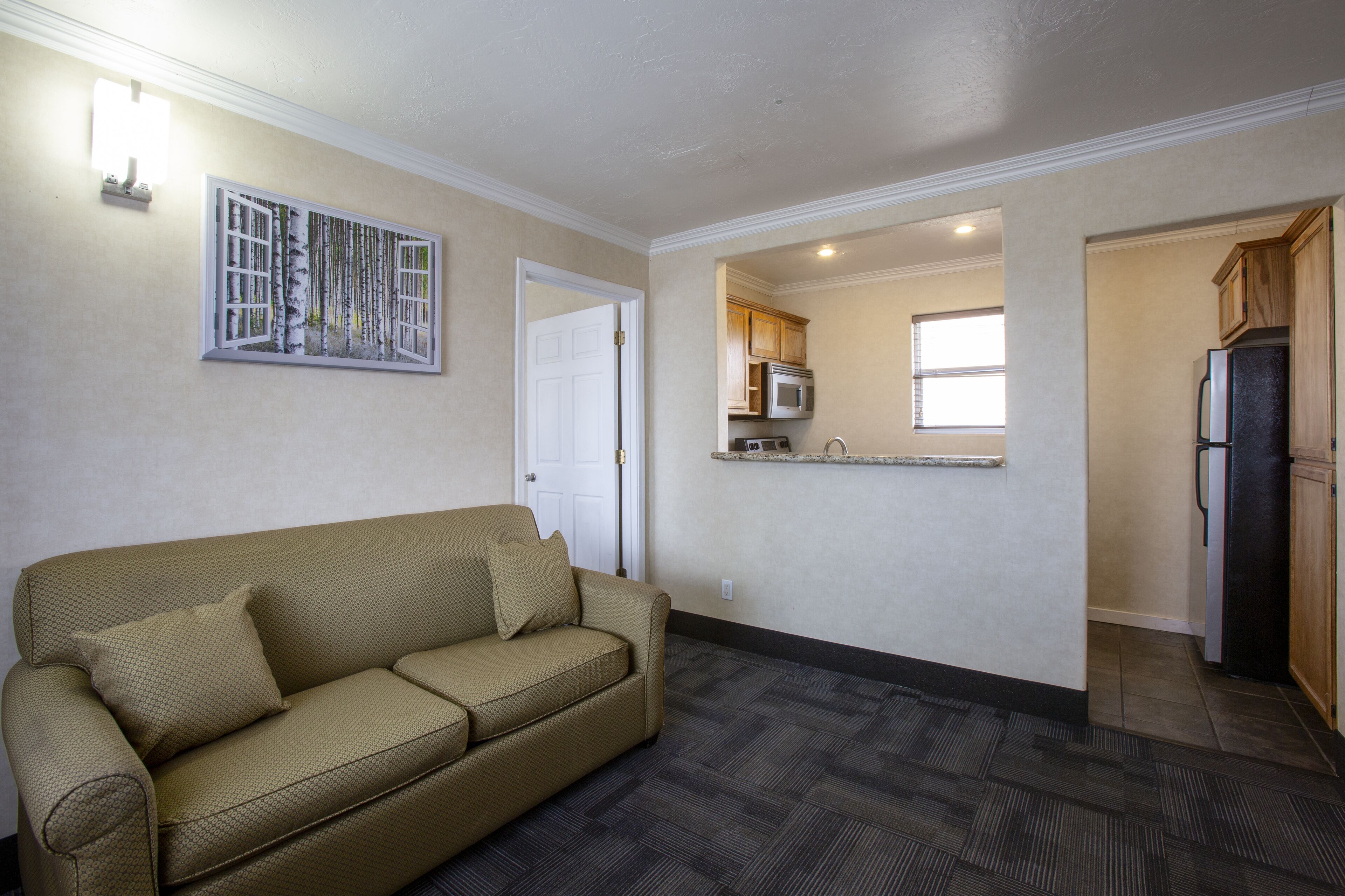 A living room with a beige sofa and a painting, a kitchen with wooden cabinets and appliances, and a door leading to another room.