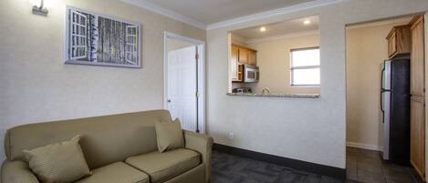 A living room with a beige sofa and a painting, a kitchen with wooden cabinets and appliances, and a door leading to another room.