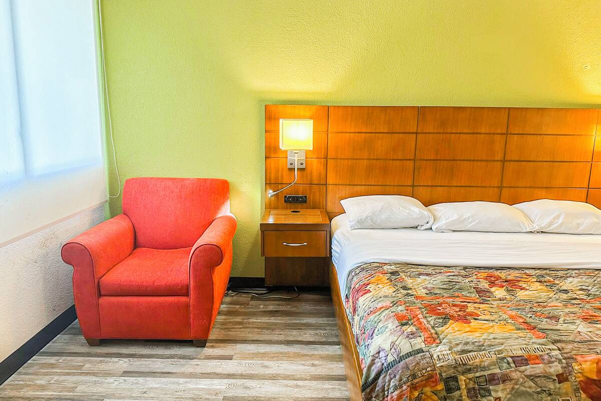 Ein Hotelzimmer mit einem hölzernen Kopfende, einem roten Sessel, einem Nachttisch mit Lampe, einem Bett mit weißer Bettwäsche und einer farbenfrohen Steude.