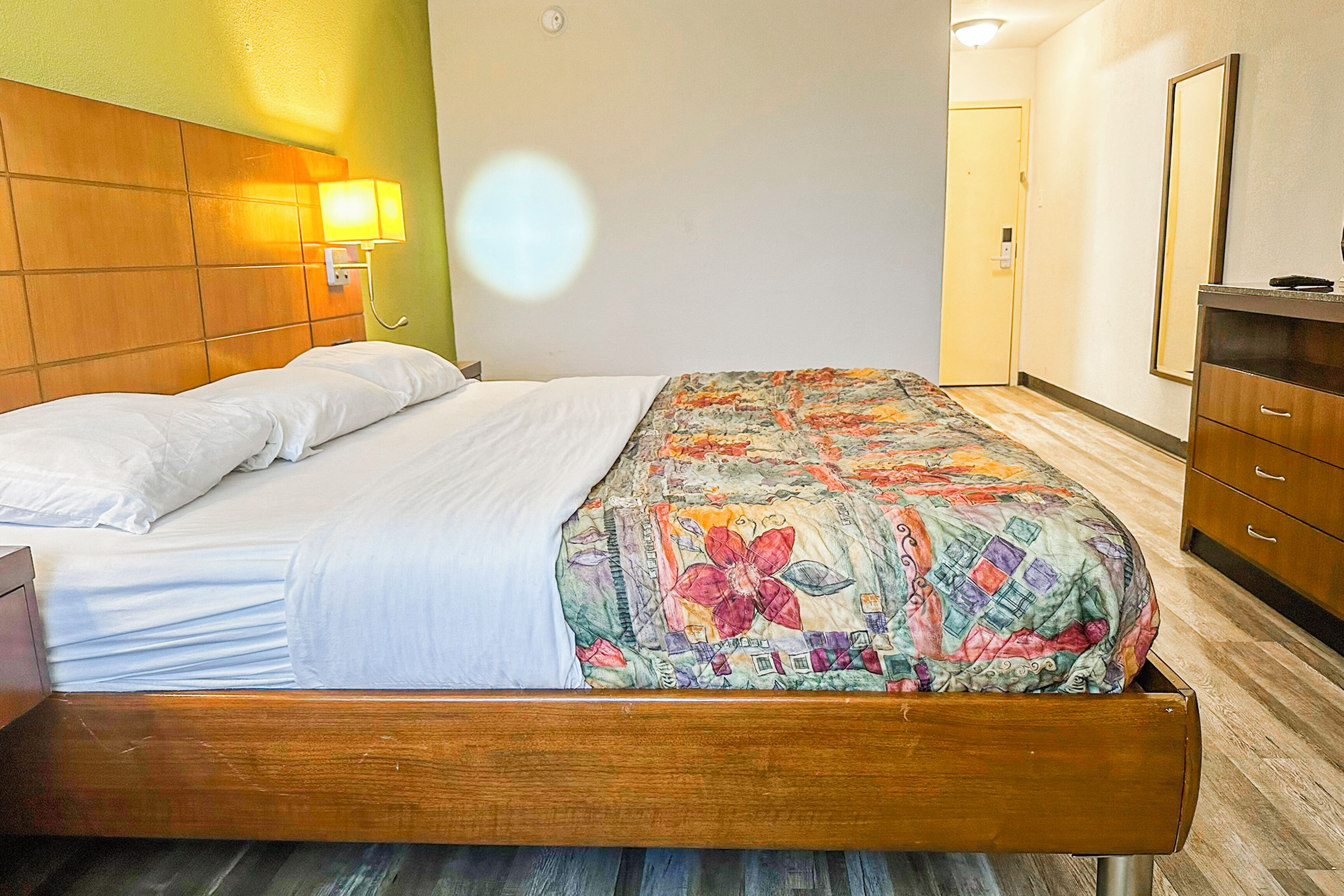 A hotel room with a wooden bed, a colorful quilt, a nightstand, a dresser, and a wall-mounted lamp.