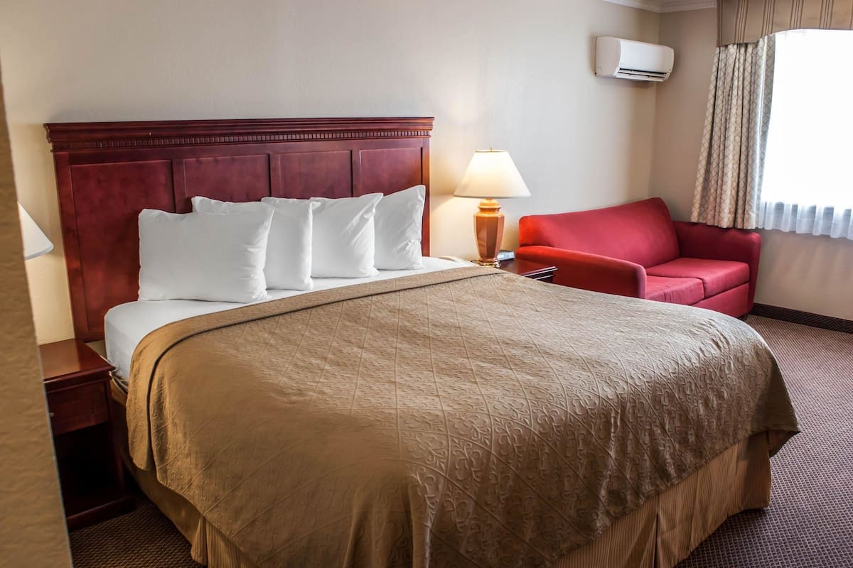 A hotel room with a large bed, a red armchair, a bedside table with a lamp, and an air conditioning unit on the wall.