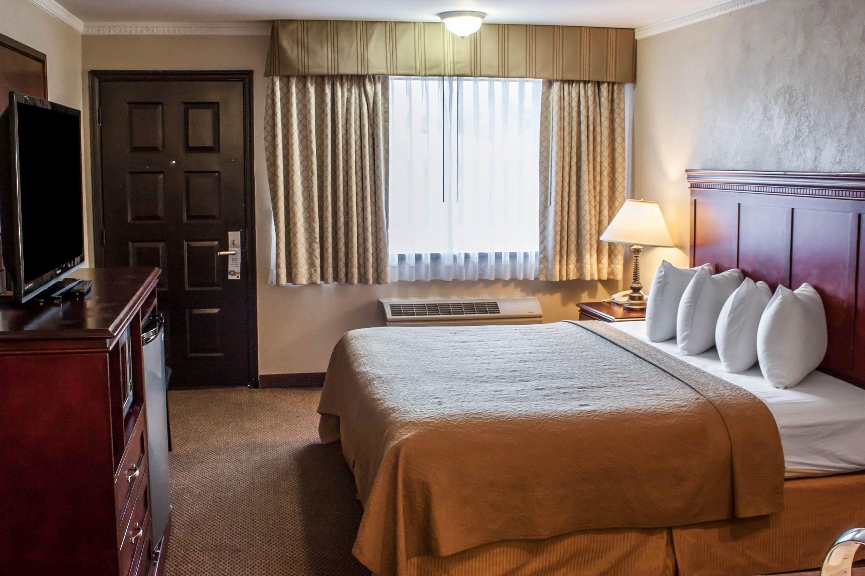 A hotel room with a bed, a television, a desk with a lamp, and a window with curtains.