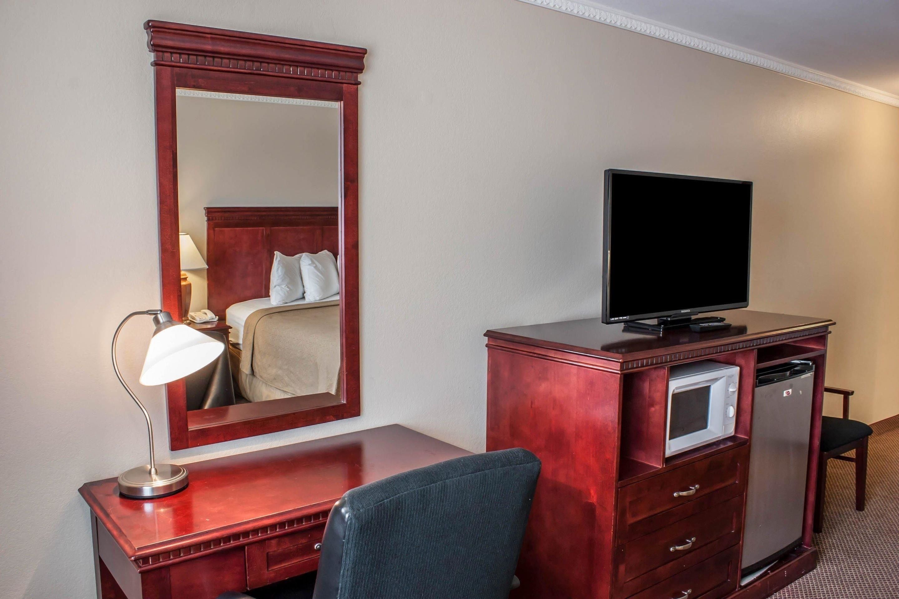 A hotel room with a wooden desk, a television, a microwave, a refrigerator, a chair, and a large mirror.