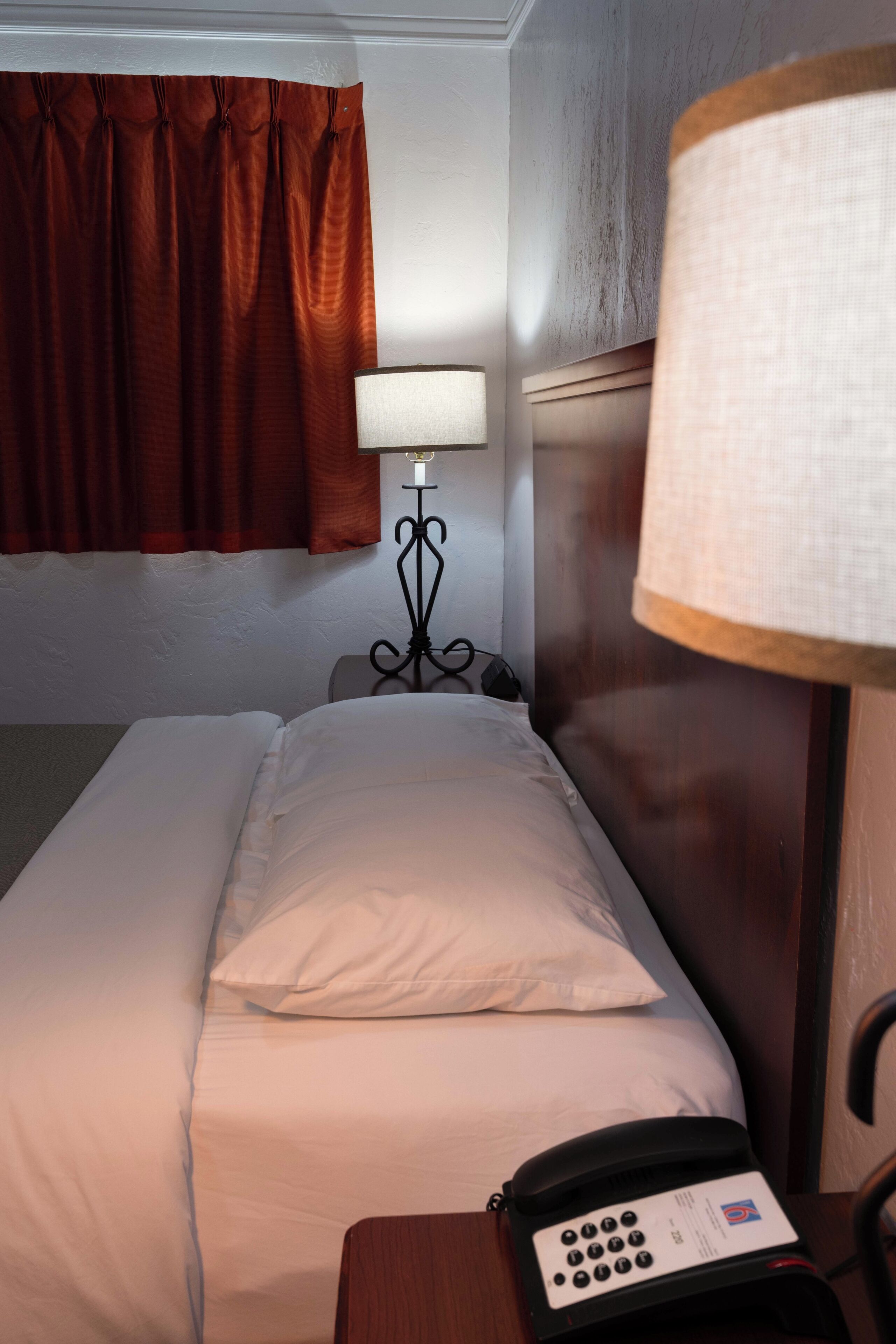 A hotel room with a bed, a bedside lamp, a telephone, and a window with curtains.