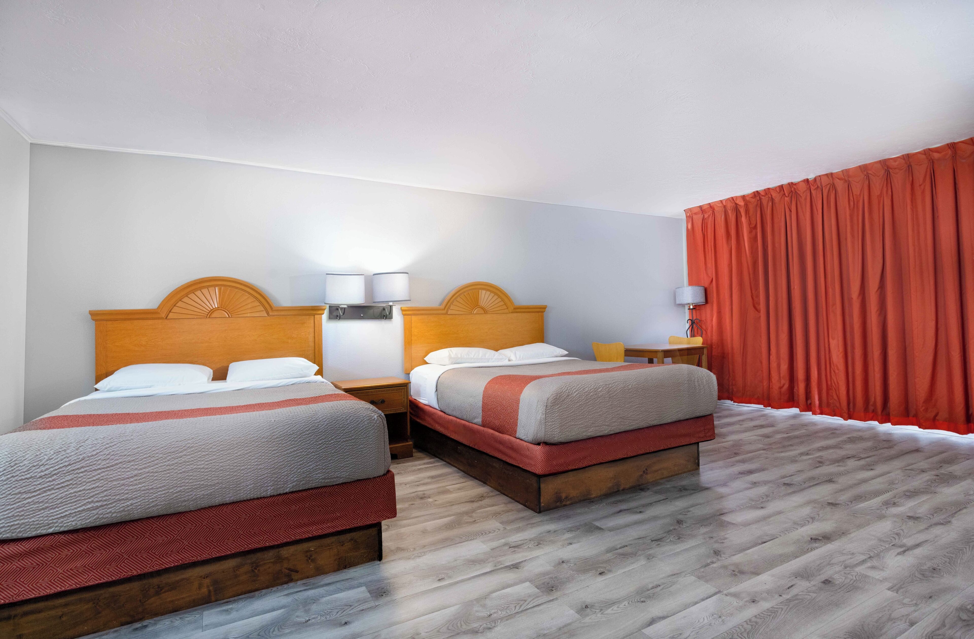 A hotel room with two beds, wooden headboards, bedside tables with lamps, and a red curtain.