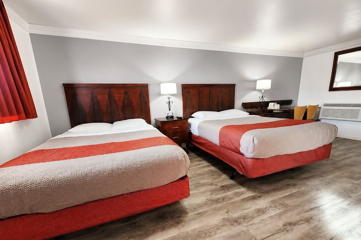 A hotel room with two beds, wooden headboards, bedside tables with lamps, a nightstand, and a window with blinds.