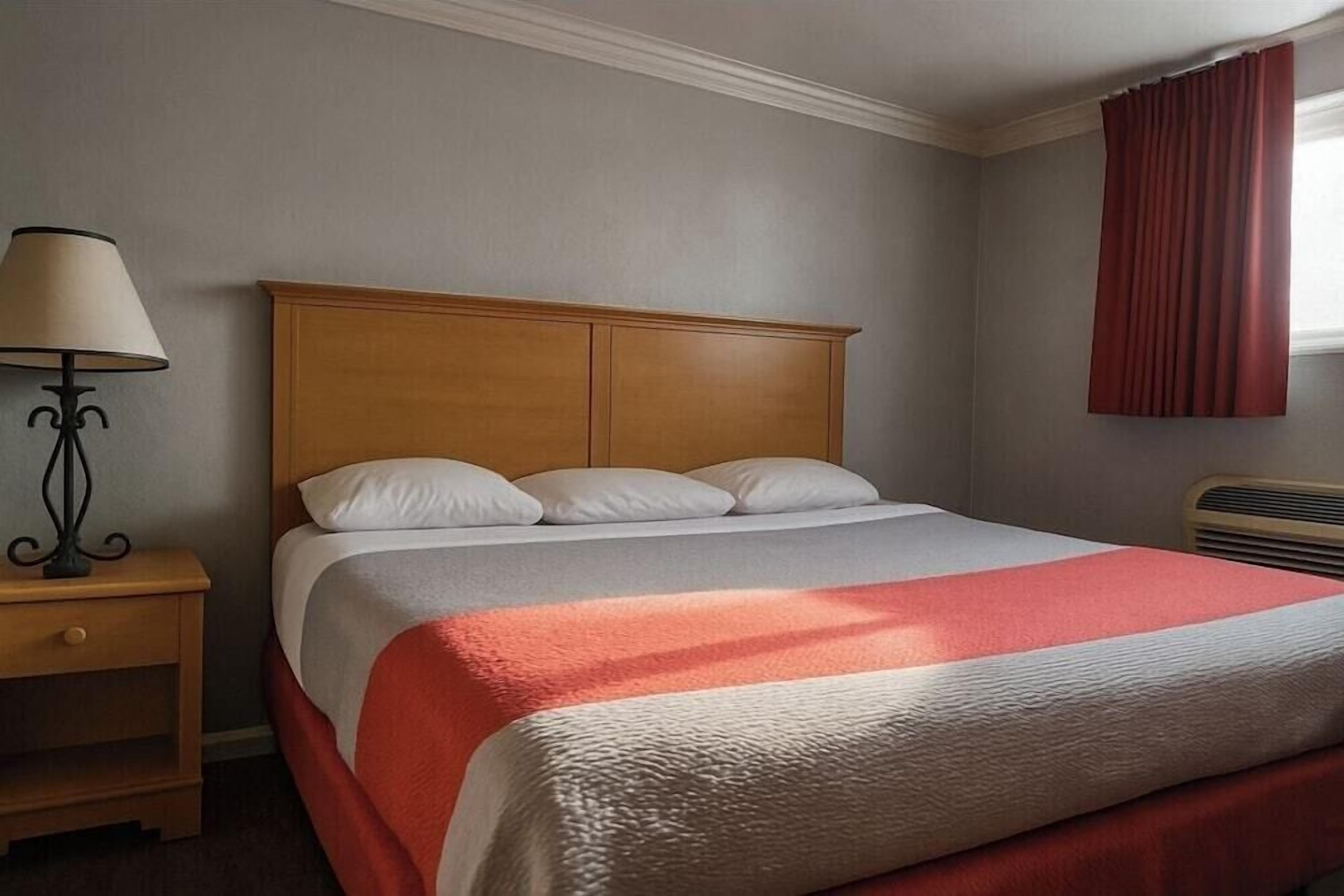 Un letto ben rifatto con poggiatesta in legno, un comodino con una lampada e una finestra con tende rosse.