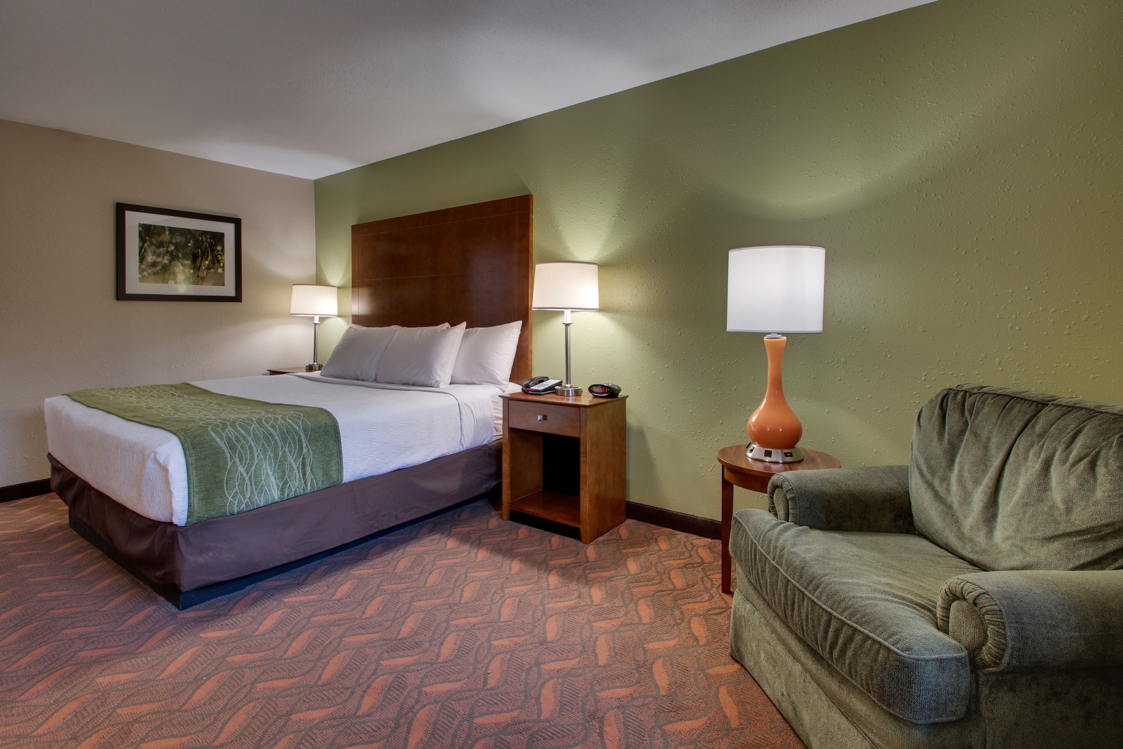 A hotel room with a bed, two bedside lamps, a nightstand, a green armchair, and a patterned carpet.