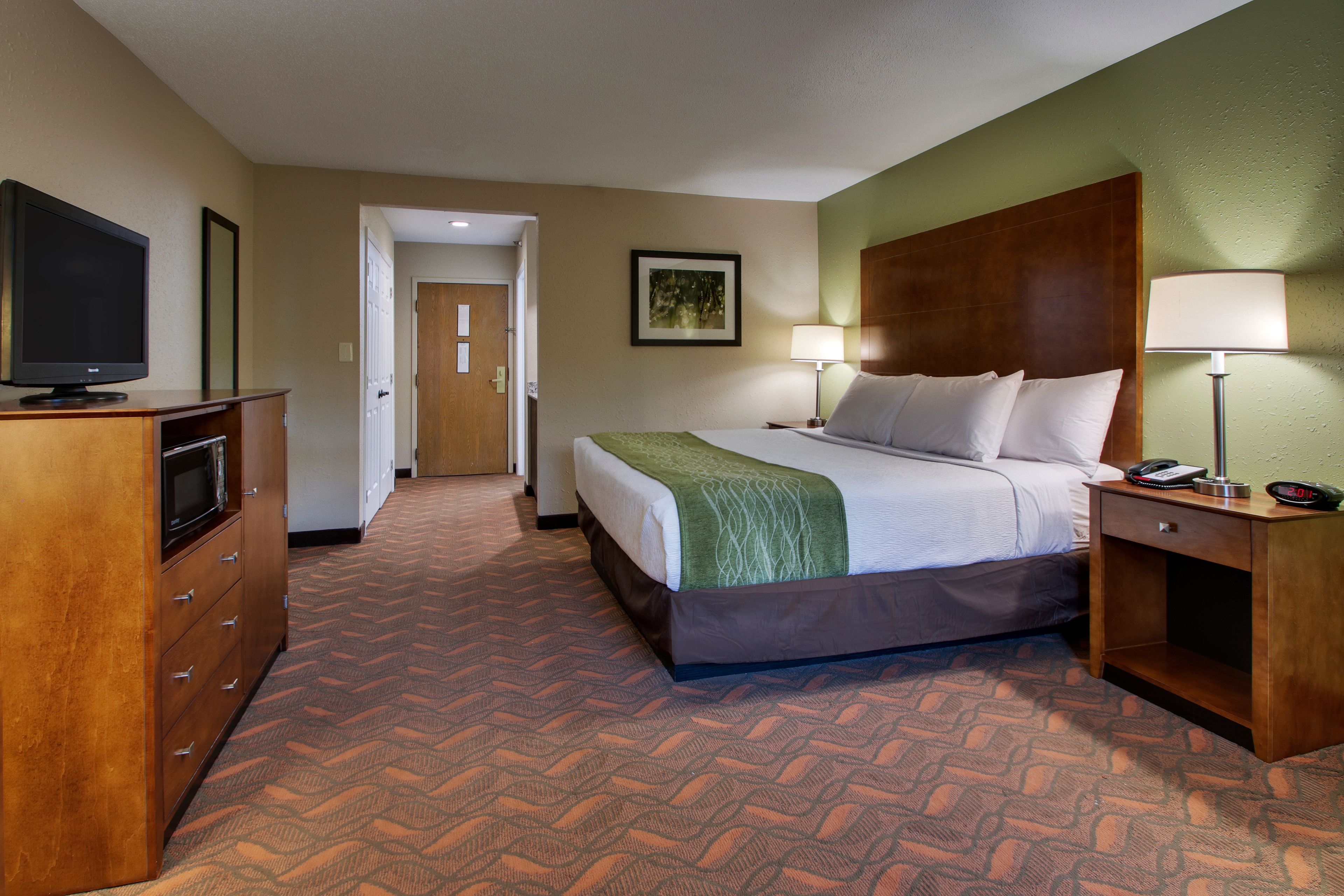 Une chambre d’hôtel avec un lit, des lampes de chevet, un bureau avec un téléphone, un téléviseur posé sur une commode et une porte donnant sur une autre pièce.