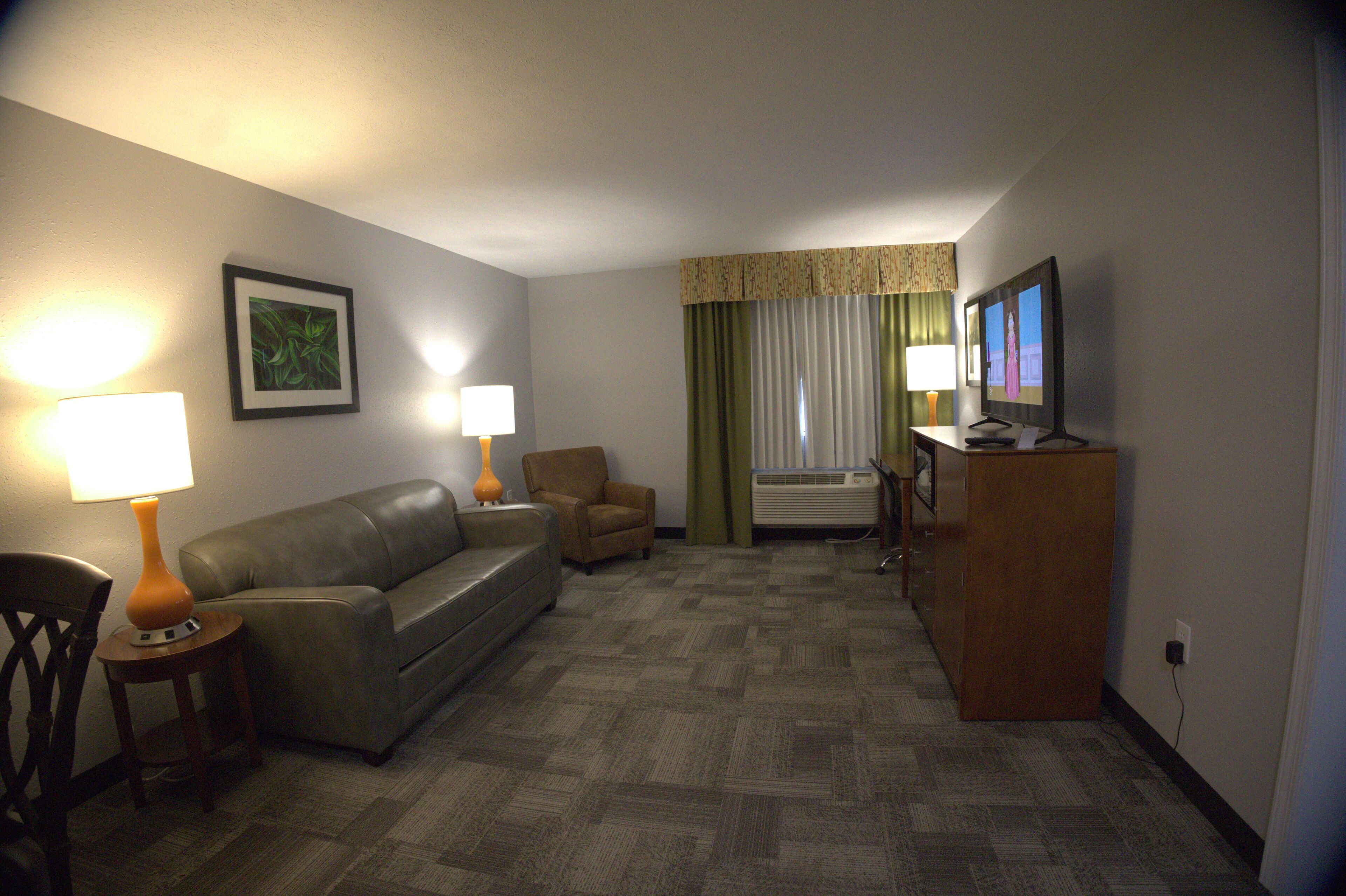 A hotel room with a sofa, armchair, TV, lamps, and a painting on the wall.