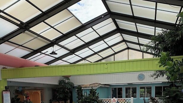An indoor swimming pool with a glass ceiling, surrounded by a green and white building, and a small seating area with chairs and tables. - Greenfield Inn and Suites Elizabethtown, KY I65 Exit 94 (Elizabethtown)