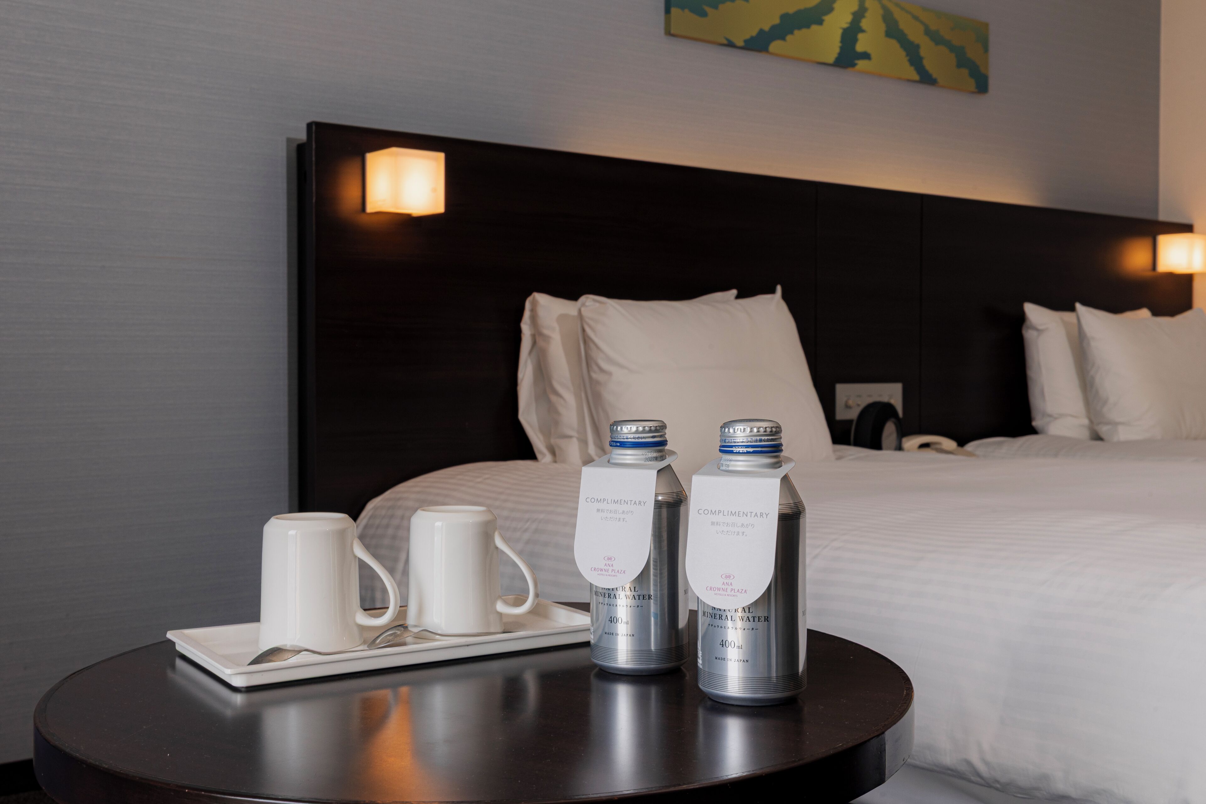 A hotel room with a neatly made bed, a bedside table with two water bottles and a tray with two white mugs.