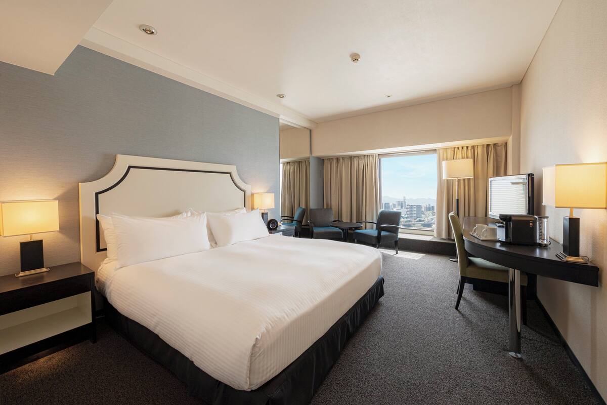 Ein Hotelzimmer mit einem großen Bett, einem Schreibtisch mit Stuhl, einem kleinen Tisch und Blick auf die Stadt durch das Fenster.